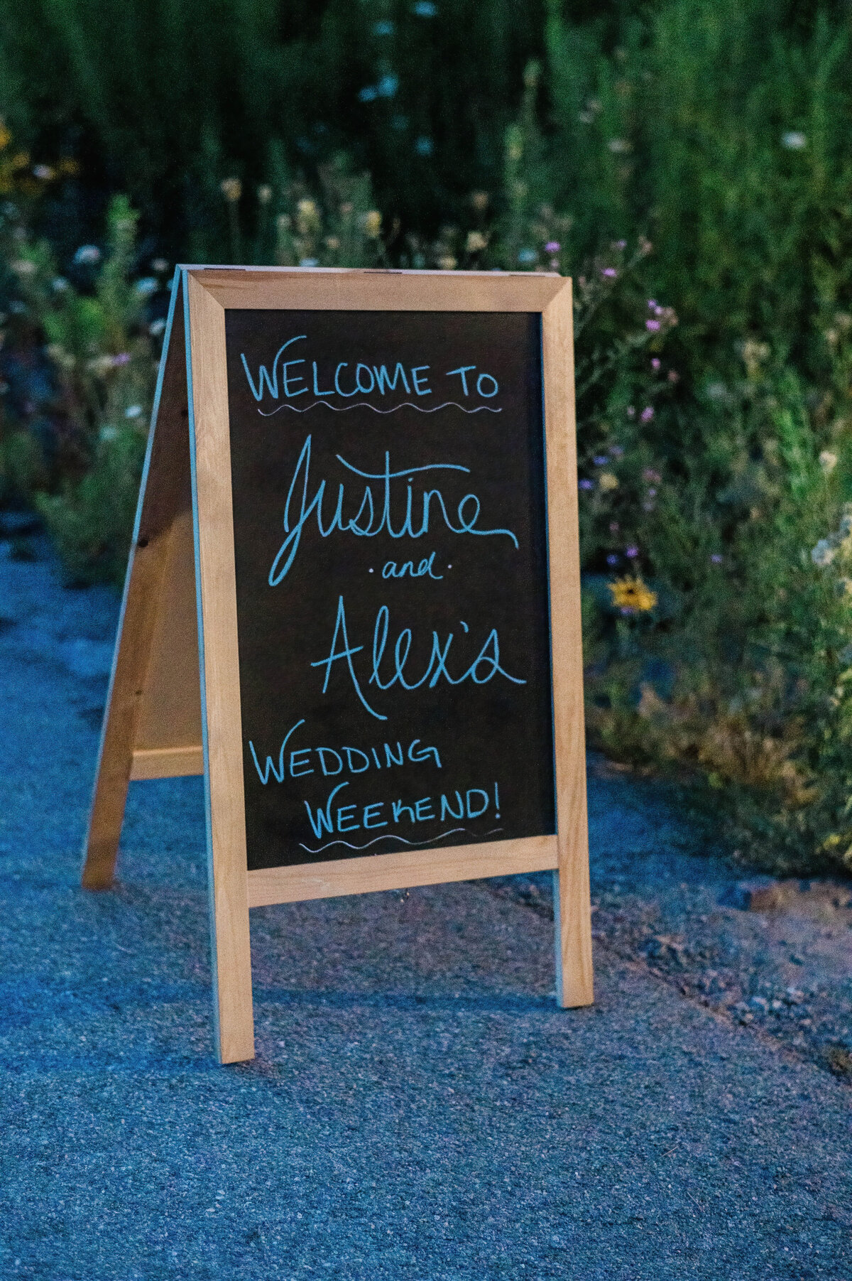 A chalkboard sign sits by a garden path, reading “Welcome to Justine and Alex’s Wedding Weekend!” in handwritten script.