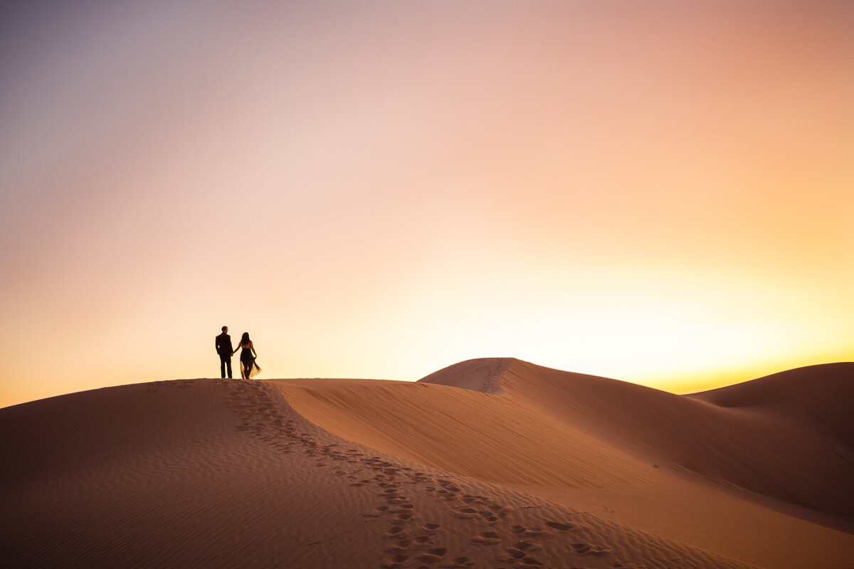 bride and groom standing at the top of a large sand dune during a colorful sunset