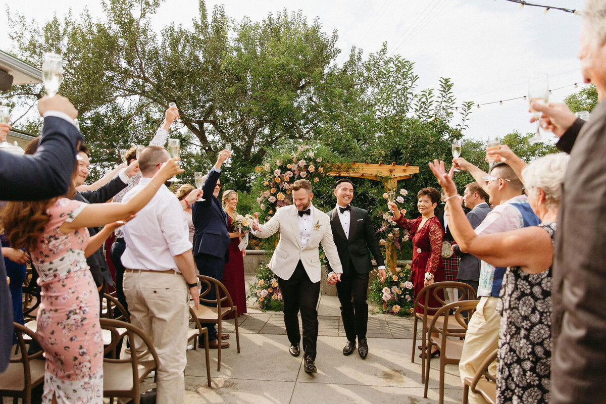 Grooms exiting ceremony, featuring  floral backdrop by The Romantiks, romantic wedding florals based in Calgary, AB & Cranbrook, BC. Featured on the Brontë Bride Vendor Guide.