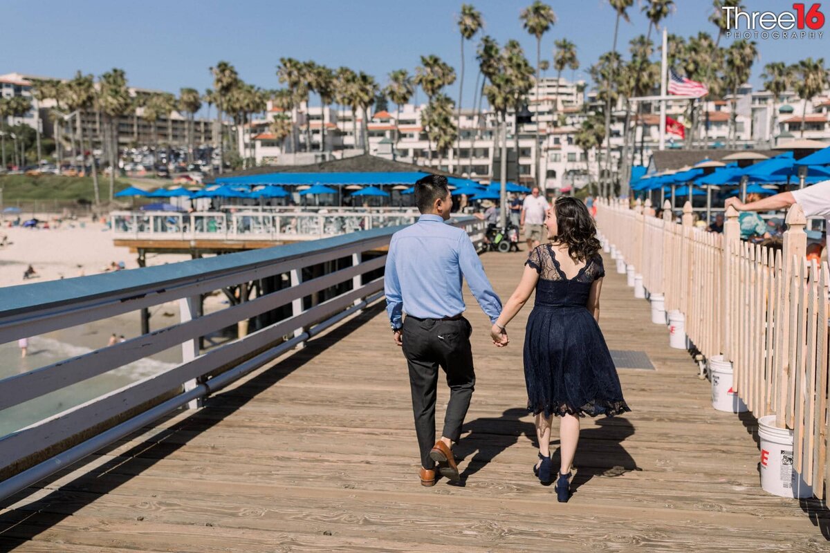 Engaged couple walk along the San Clemente Pier with the Pier Bowl in front of them