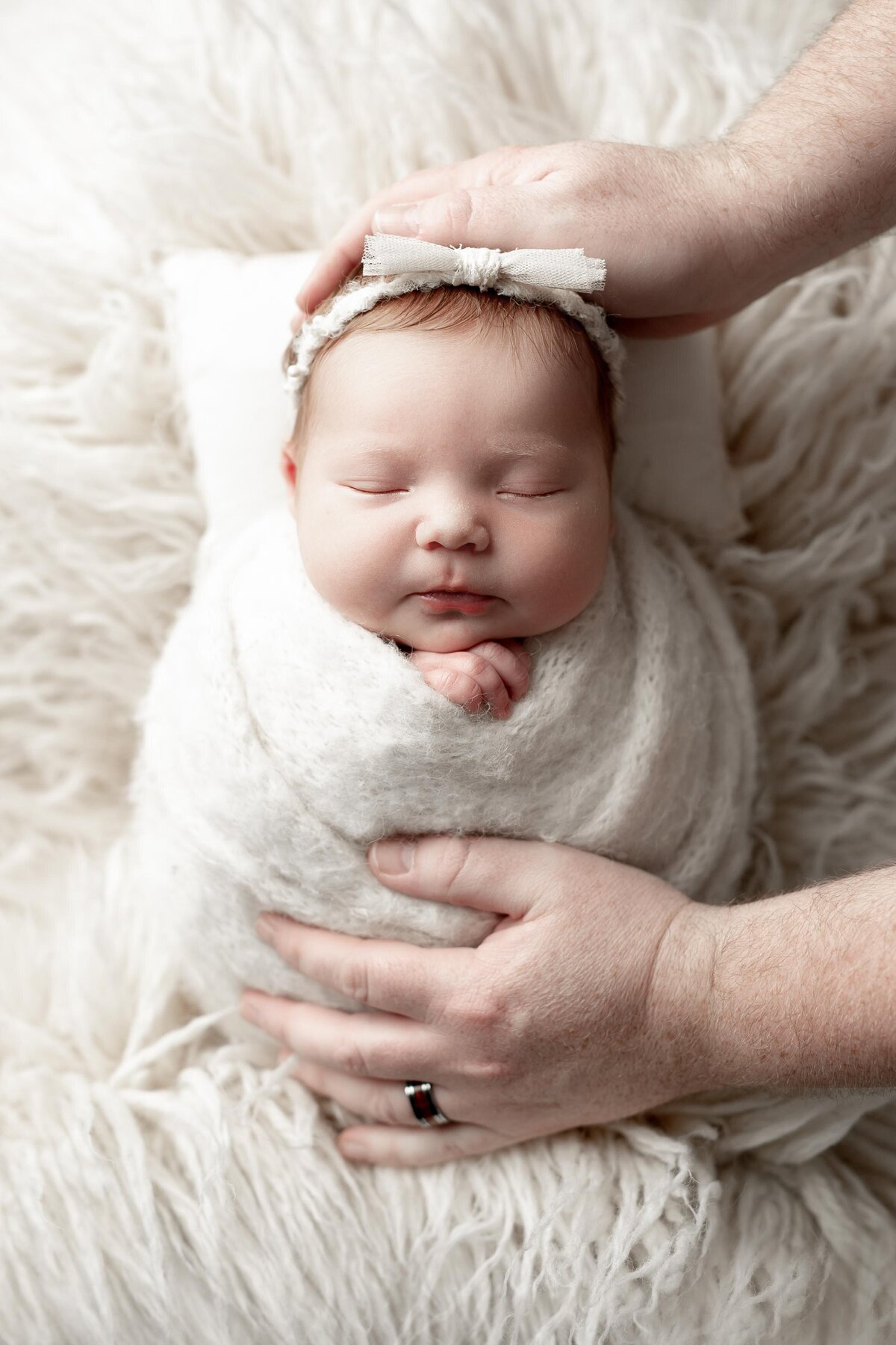Studio newborn photography - sleeping baby swaddled in cream wrap with delicate bow headband. Dad's hands are gently resting on the bottom of the swaddle and the top of her head to show the scale of how small the baby is.