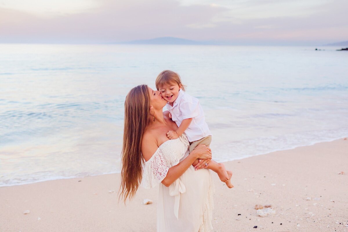 Sunrise portrait of a young boy being lifted and kissed by his mother on Maui who wears a white off-shoulder dress at the beach