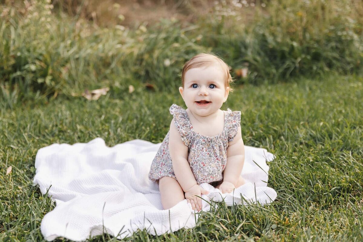 Young baby girl sitting on white blanket surrounded by lush greenery during Toronto Family Portrait session. 