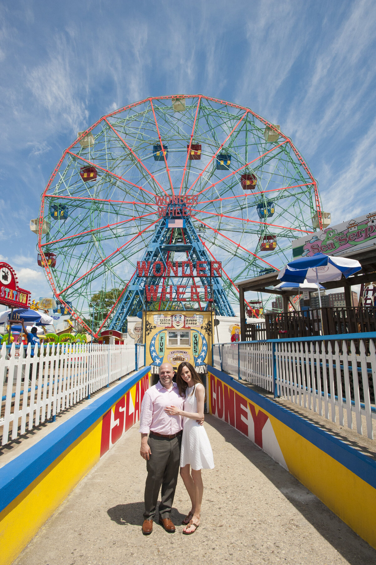 A couple standing side by side smiling in front of a ferris wheel on Coney Island 