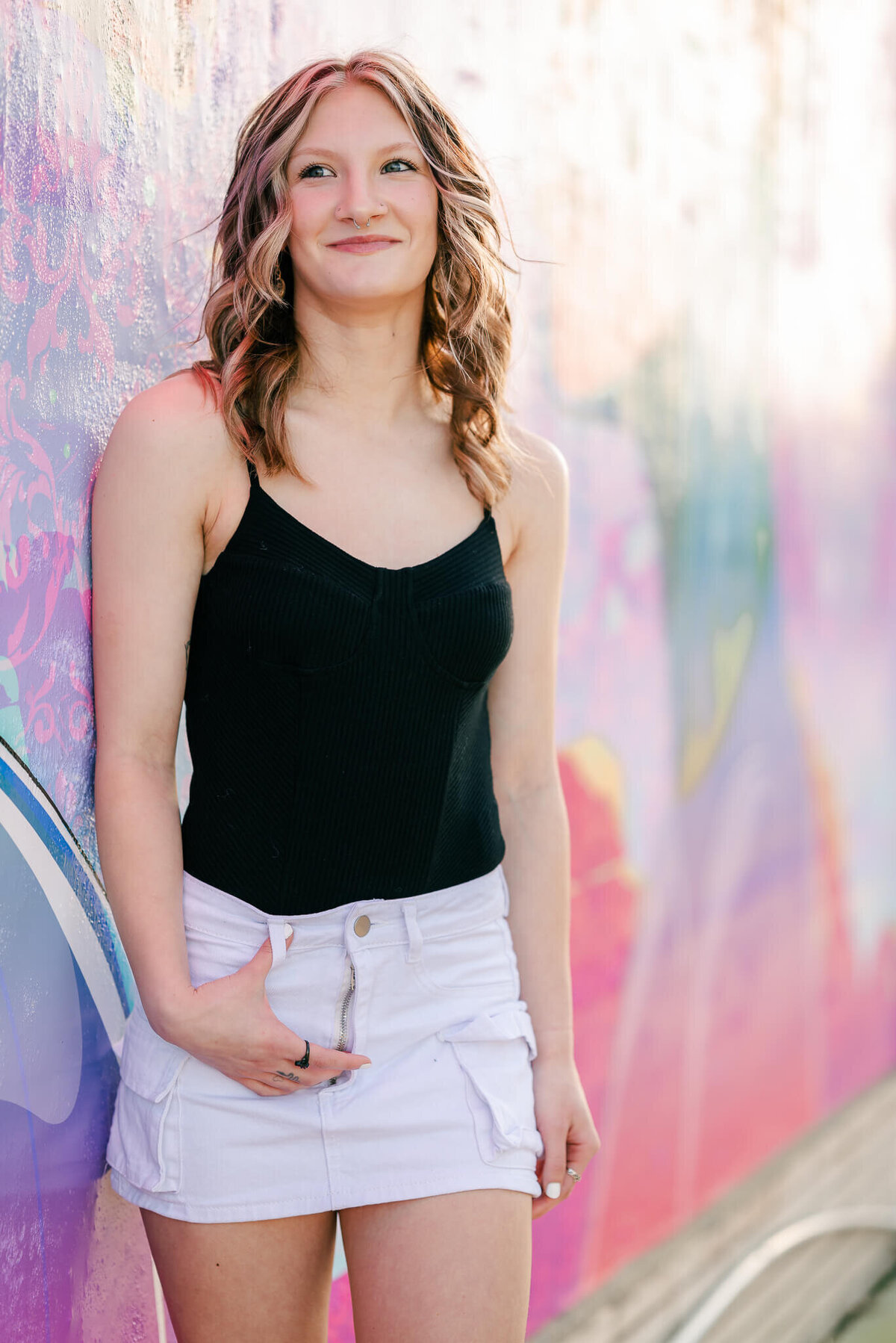 A high school senior leans against a mural in Norfolk's Neon District. She is holding back a laugh white she hooks her hand in her white denim skirt's belt loop.