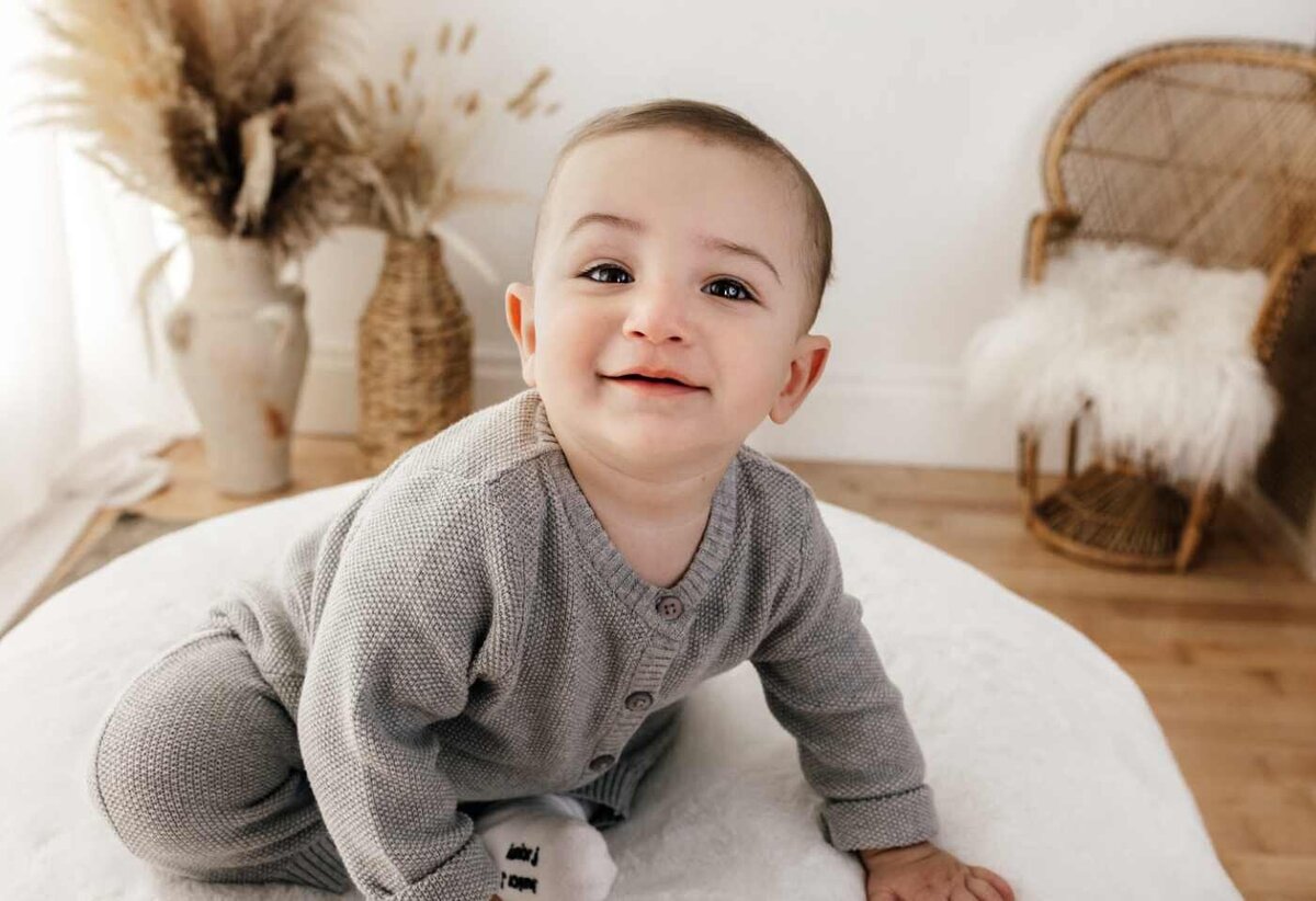 Ruua Hamdan Photography (UK): A smiling baby waering a baby suit sitting on a white blanket.