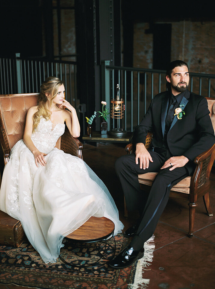 Bride and groom wearing a black tuxedo and white wedding gown sit on blush-colored chairs.