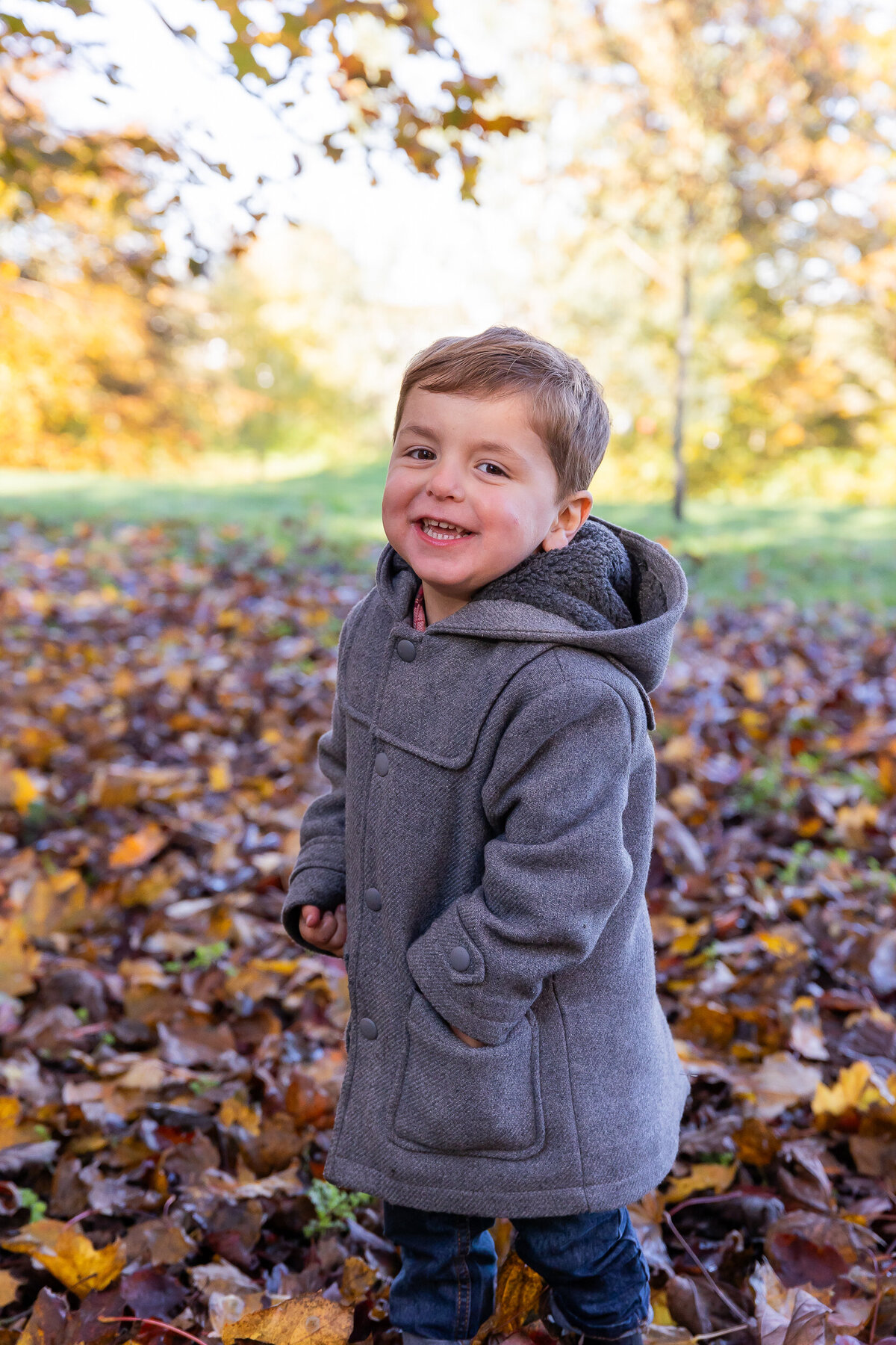 A young boy in a gray coat smiles joyfully while standing in a park covered with autumn leaves.