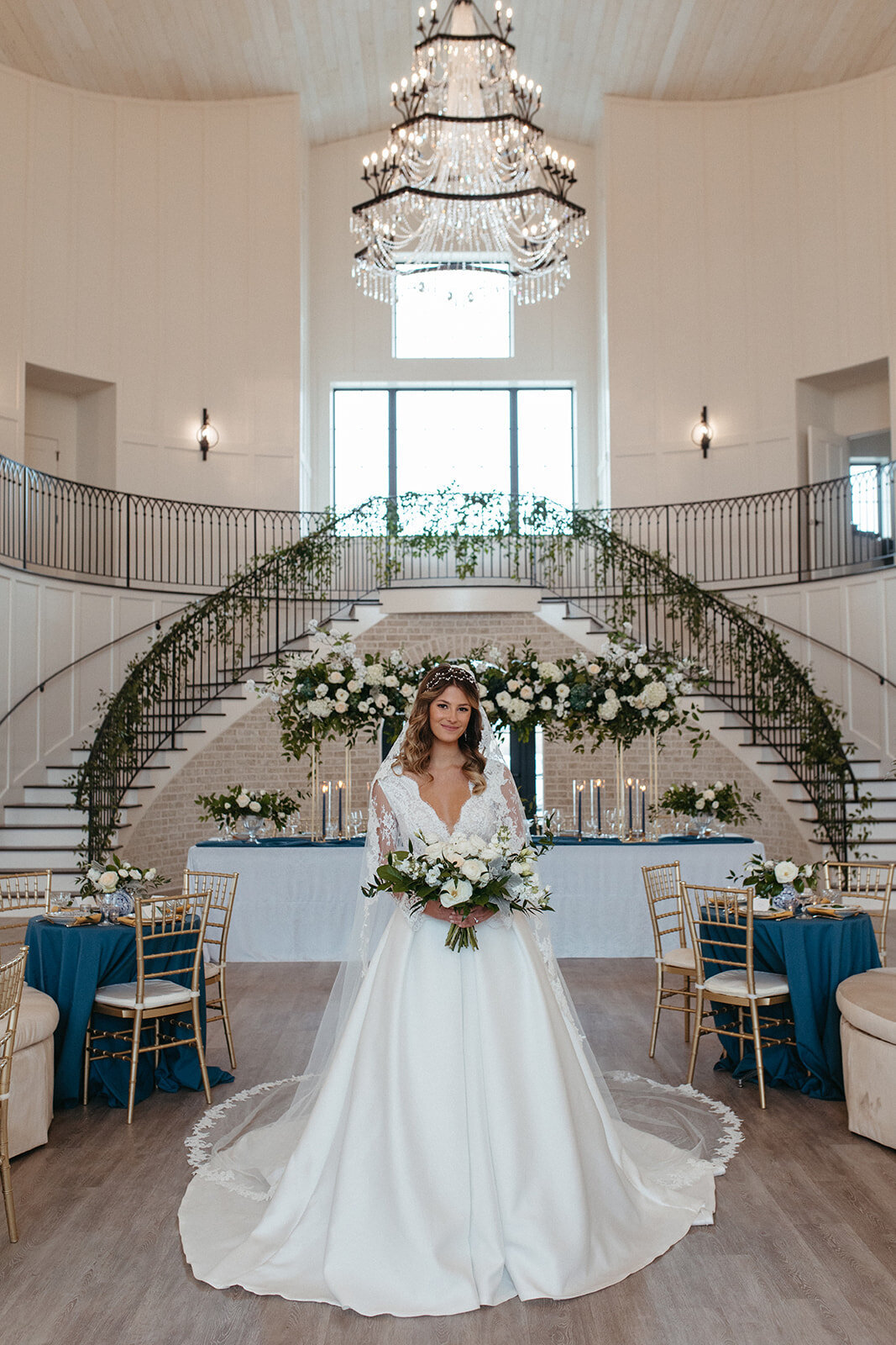 Bride wearing a white wedding gown and veil holding a white bouquet in the middle of a banquet room.