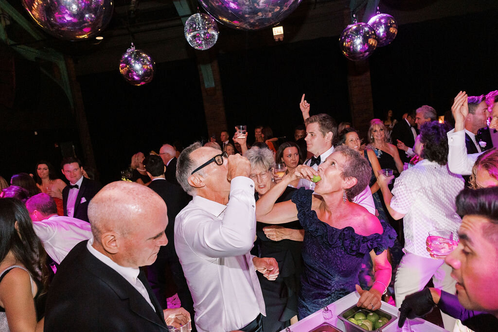 Wedding guests enthusiastically take celebratory shots together beneath reflective disco balls, embracing the party atmosphere.