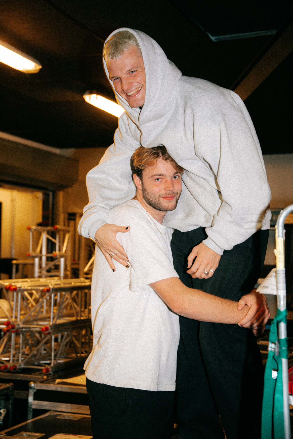 Sam Hofman is shown in an embrace with a friend at a festival, both smiling warmly. Sam, shirtless and covered in tattoos, hugs his friend, who is dressed in a white crop top and black skirt. The festival setting in the background adds to the joyful and celebratory mood of the image. (Photo by Bo Eppink)