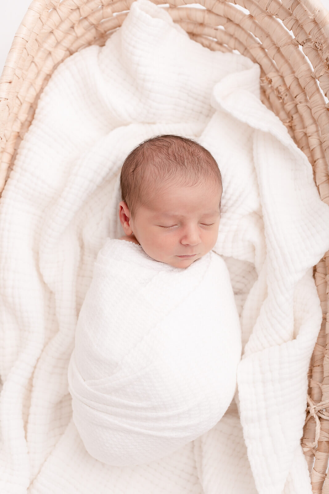 Baby wrapped in white and laying on white textured blankets inside a moses basket. The image is shot from above and only shows parts of the moses basket.