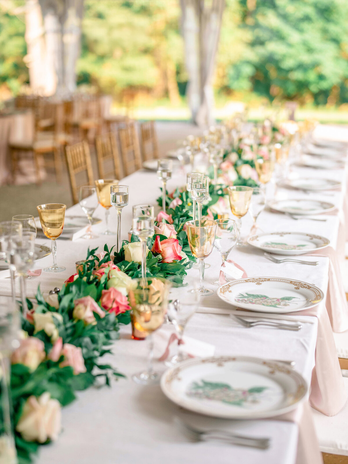 A beautifully set outdoor banquet table with elegant white tablecloths, gold-rimmed glassware, and floral centerpieces featuring pink and white roses. Plates have a floral design, and there are trees visible in the blurred background.