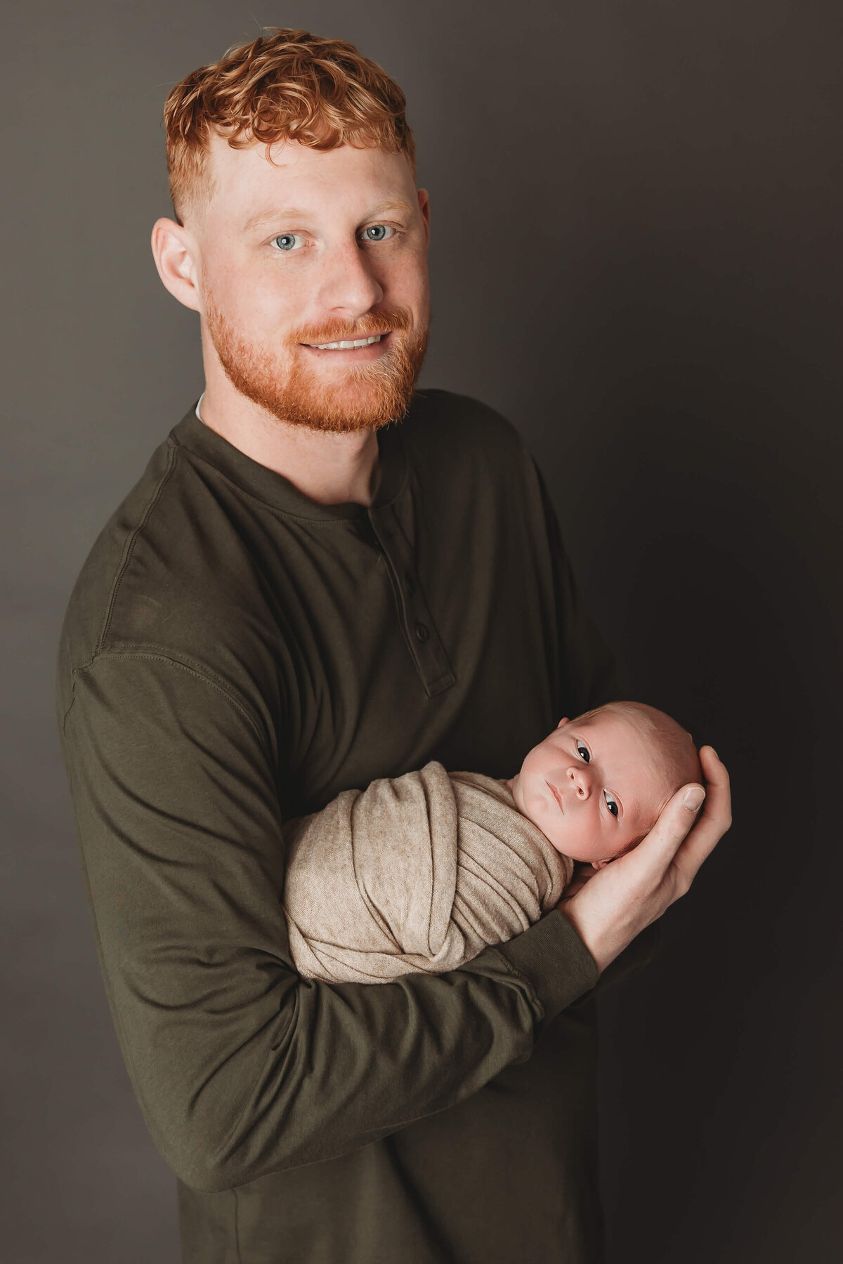 dad proudly holding his newborn son baby is a wake and they are both looking at the camera 