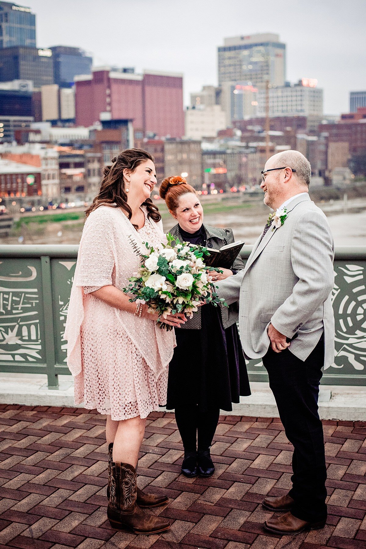 The bride and officiant bother smile at the groom during the wedding ceremony on the pedestrian bridge in Nashville. The Nashville skyline is behind them. The bride is wearing a flowing blush  gauze  dress that comes to her knees with brown cowboy boots. She is holding a large white bouquet of flowers with greenery. The officiant is wearing a black dress and she is holding a leather bound book. Her red hair is up in a structured bun. The groom is wearing black pants and a light gray jacket. His hands are in his pockets.