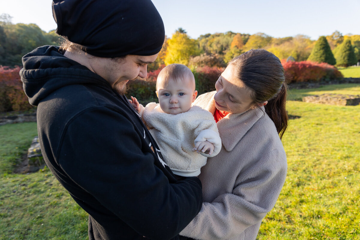 A young family with a baby standing outdoors in a sunlit park during autumn. the parents look lovingly at their child.