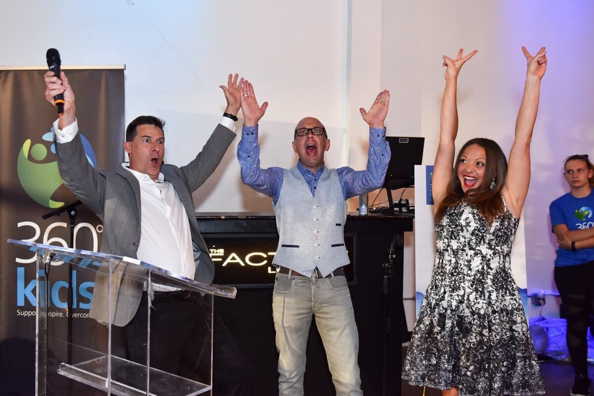 Three charity event speakers celebrating with enthusiastic cheers on stage during a Toronto event, captured by a professional event photographer.
