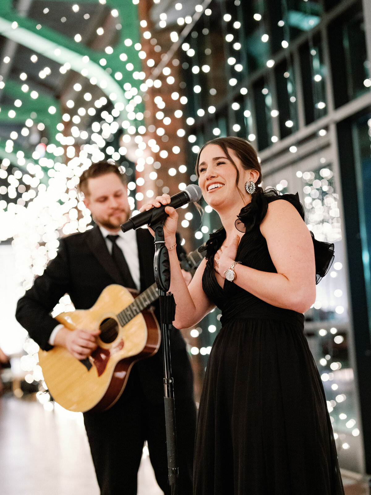A close up of a woman speaking into the microphone and guitar player in the background.  