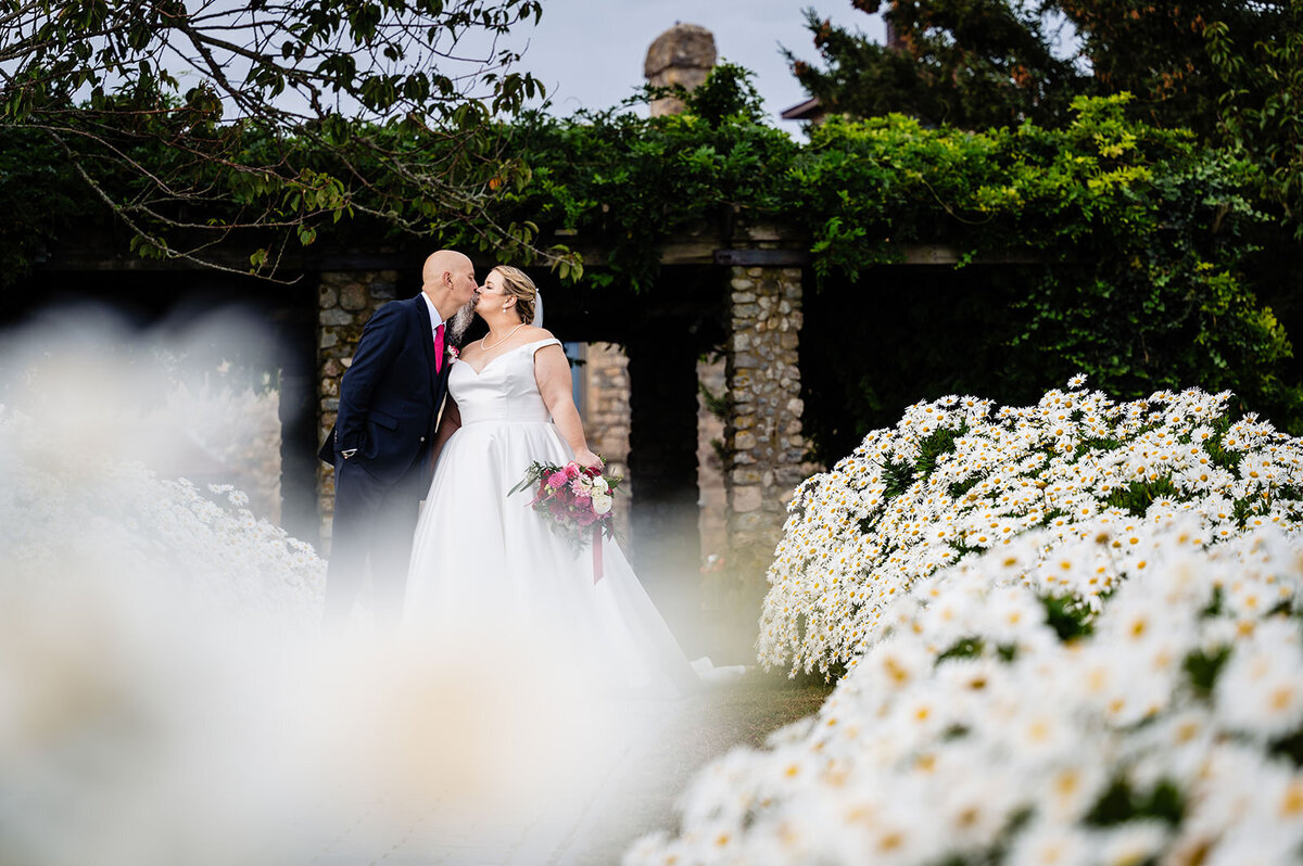 A bride and groom sharing a kiss on a garden path, surrounded by white flowers with a stone arch in the background