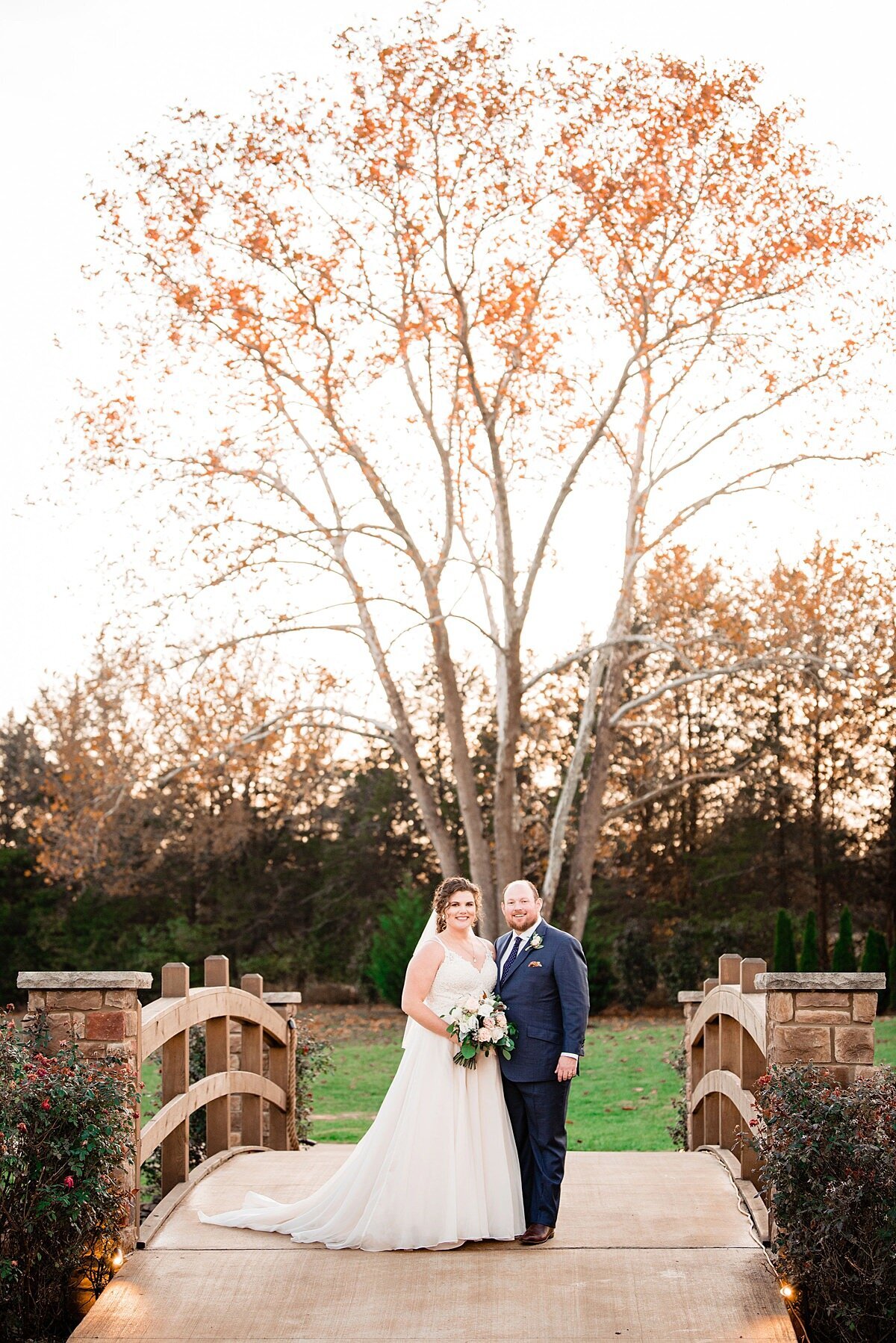 The bride, wearing a long, a-line, lace wedding dress and veil holding a white, ivory and blush bouquet standing beside the groom dressed in a navy blue suit on a bridge passing over a creek at Sycamore Farms