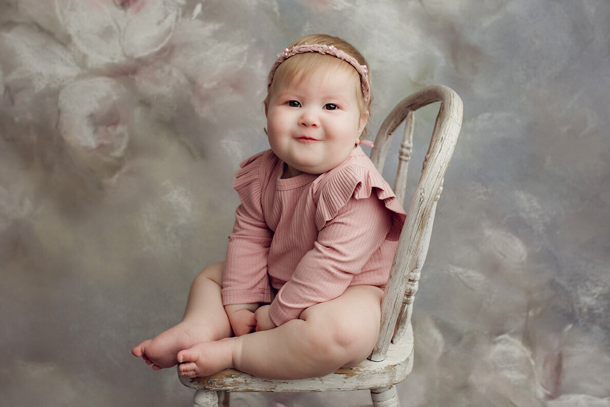 a one year old girl sitting in a white chair with a pink frilly romper