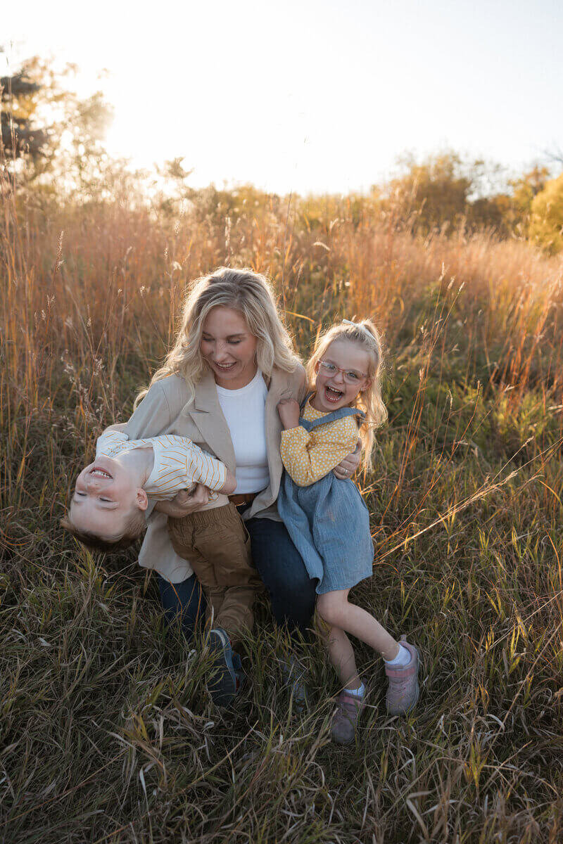 A playful outdoor family photo featuring a mother sitting in tall grass, holding her laughing son upside down while her daughter smiles beside her.