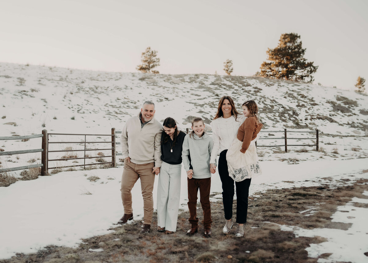A family of five walks on a trail, surrounded by beautiful snowy hills and a gorgeous old fence, while dressed fashionably in Brown's and creams to coordinate with the winter backdrop.
