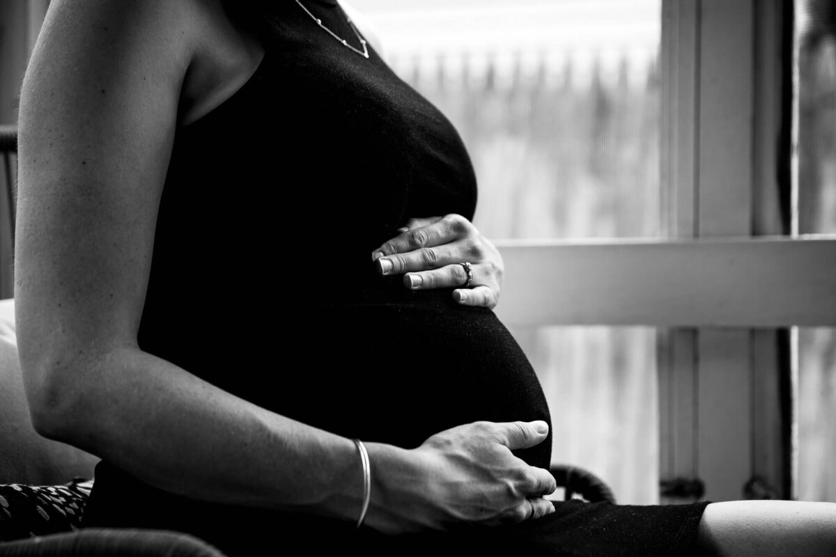 Black and white fine art portrait of a pregnant woman's stomach, taken in her sun porch. She's wearing a sleeveless black dress and has a silver bangle on her right wrist, one hand above her tummy and one below.