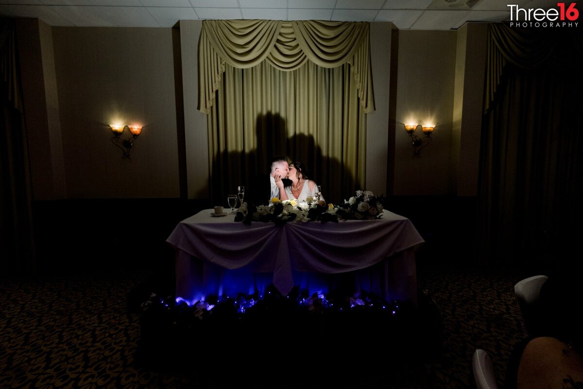 Newly married couple kiss while sitting at the sweethearts table in the dark