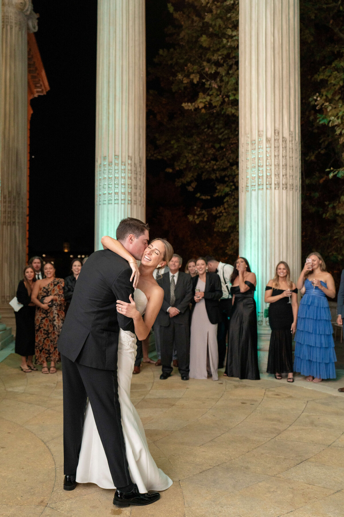 A couple shares a joyful dance in front of tall pillars during an outdoor evening event. The bride wears a strapless white gown, and the groom is in a black suit. Guests watch and smile, some dressed in formal attire. The setting is softly lit.