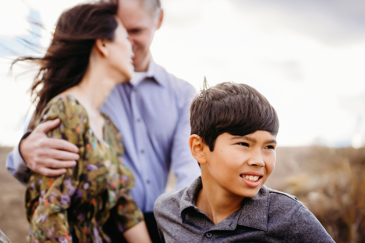 Family photo session at Standley Lake in Westminster Colorado.  Mom and Dad hug while boy looks away in the wind on chilly afternoon.