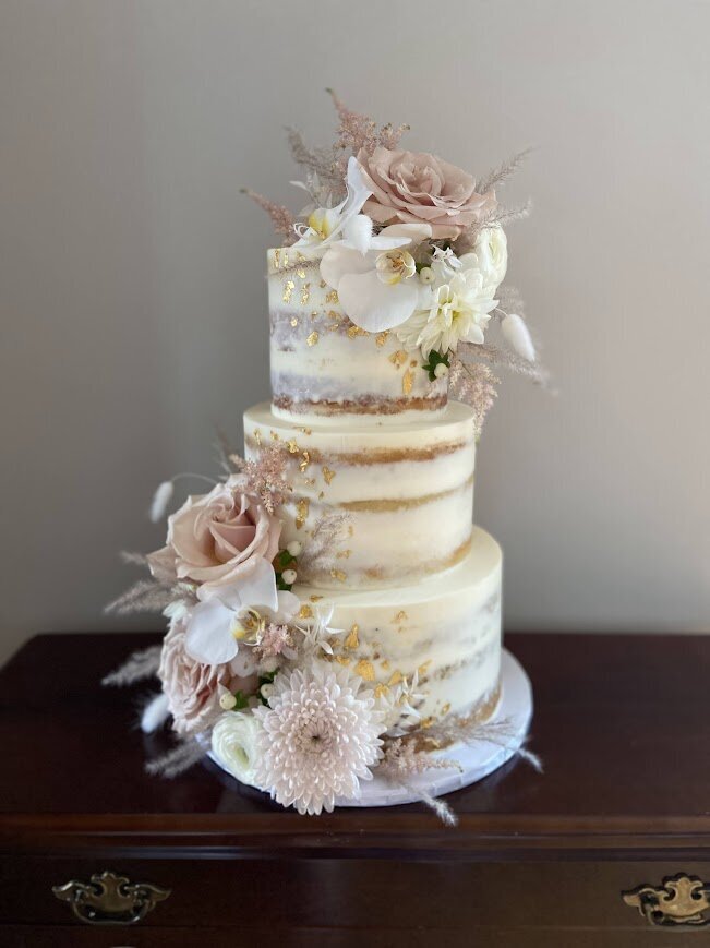 Photo is of a three tier cake with naked style frosting. Decorated with large clusters of fresh flowers in pinks and whites, with gold leaf.