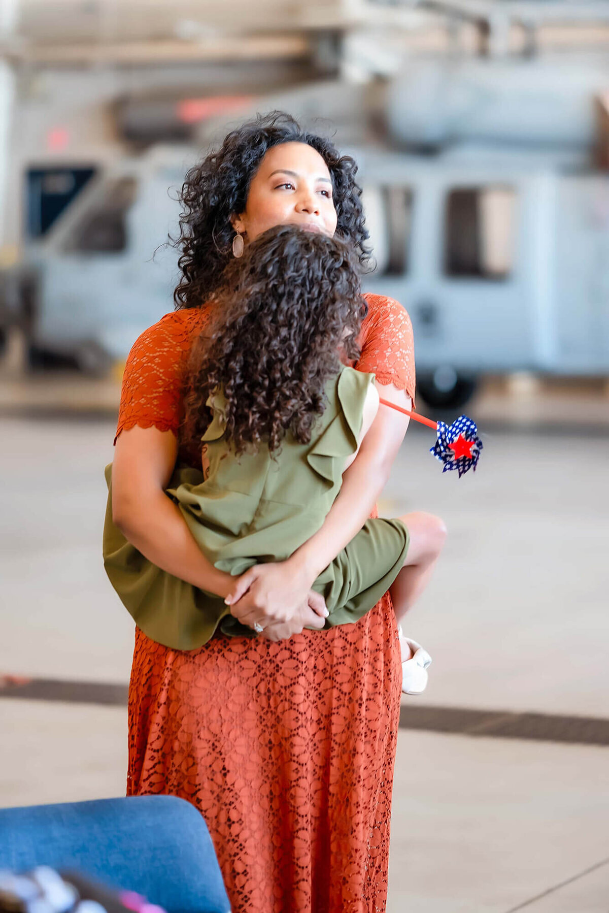 A woman in an orange dress holds her young daughter in her arms and looks out at the flight deck. There is a helicopter in the hangar behind them.