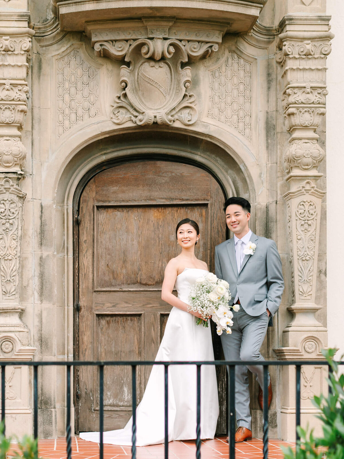 Bride in a strapless white gown holds a bouquet, standing next to a groom in a light gray suit. Both smile warmly in front of an ornate wooden door.