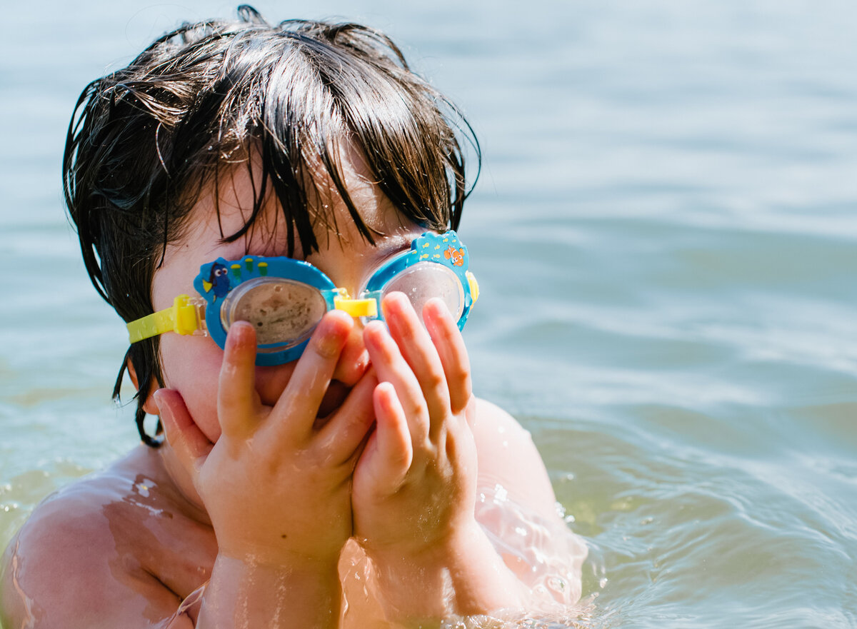 A small child in the water with goggles on as they cover their face with their hands.