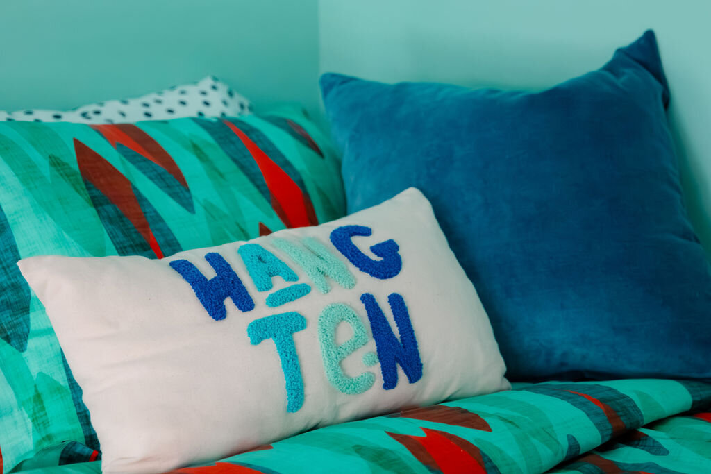 A close-up of a bed featuring colorful bedding and pillows. A teal comforter with abstract orange and darker teal shapes is layered with a white pillowcase that has black polka dots. Two decorative pillows rest against the headboard: a rectangular white pillow with the embroidered words "HANG TEN" in blue and teal lettering, and a larger, solid teal velvet pillow. The light turquoise wall serves as a backdrop. The lighting is soft and even.