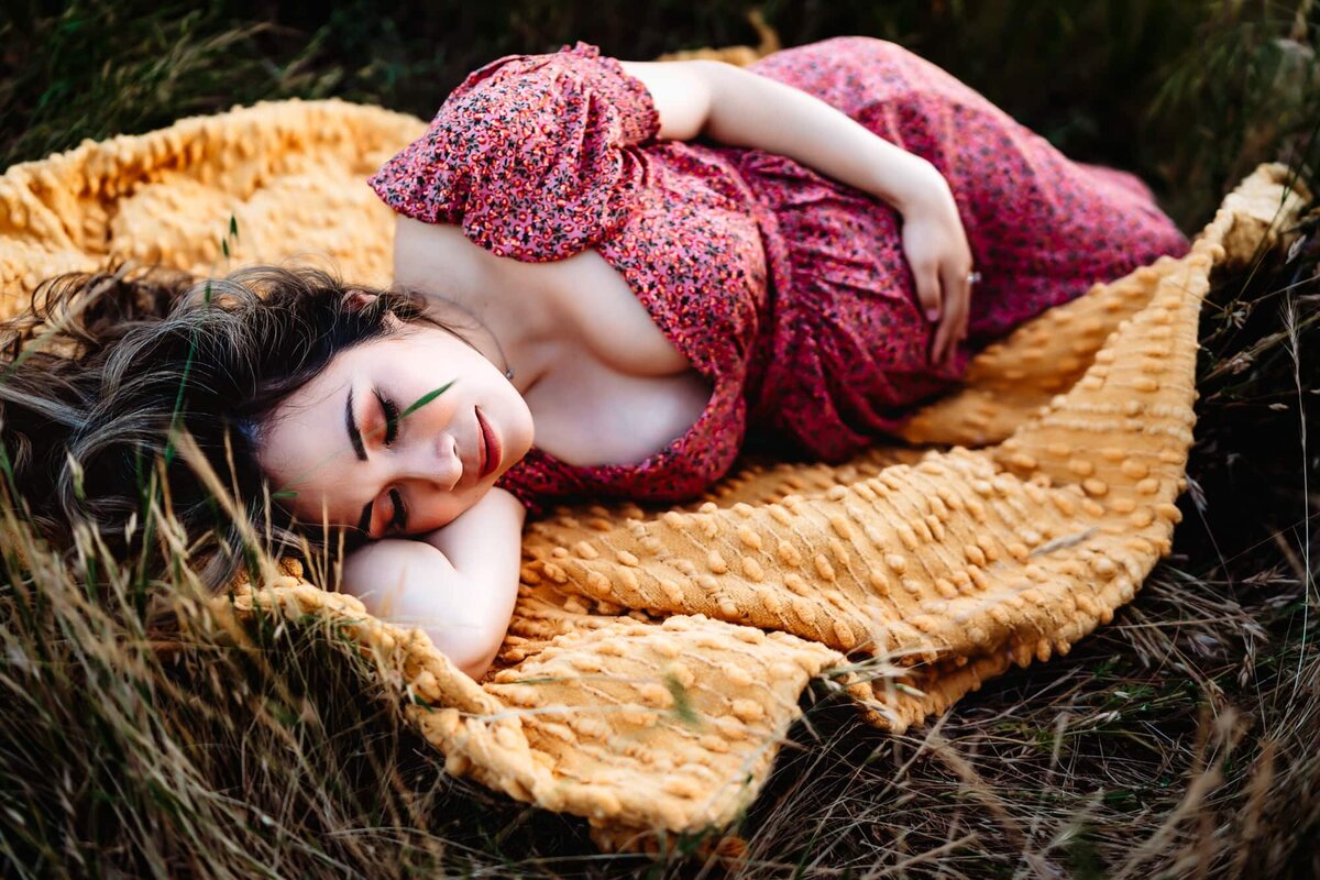 A pregnant woman wearing a pink dress lying down on a yellow blanket in a field, closing her eyes and holding her belly during a San Diego lifestyle maternity photo session by Love Michelle Photography