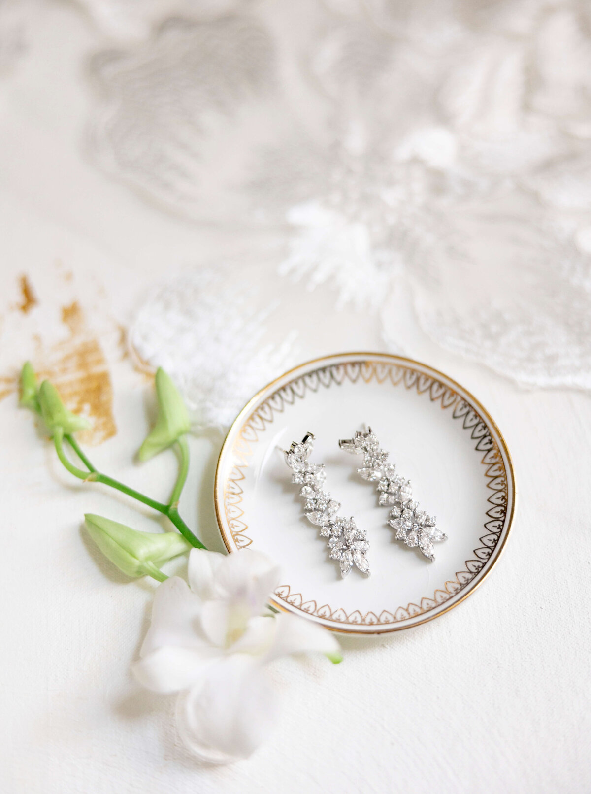 Elegant silver earrings with floral design rest on a decorative white dish with a gold rim. Beside the dish, delicate white flowers and green stems are arranged on a textured white surface.