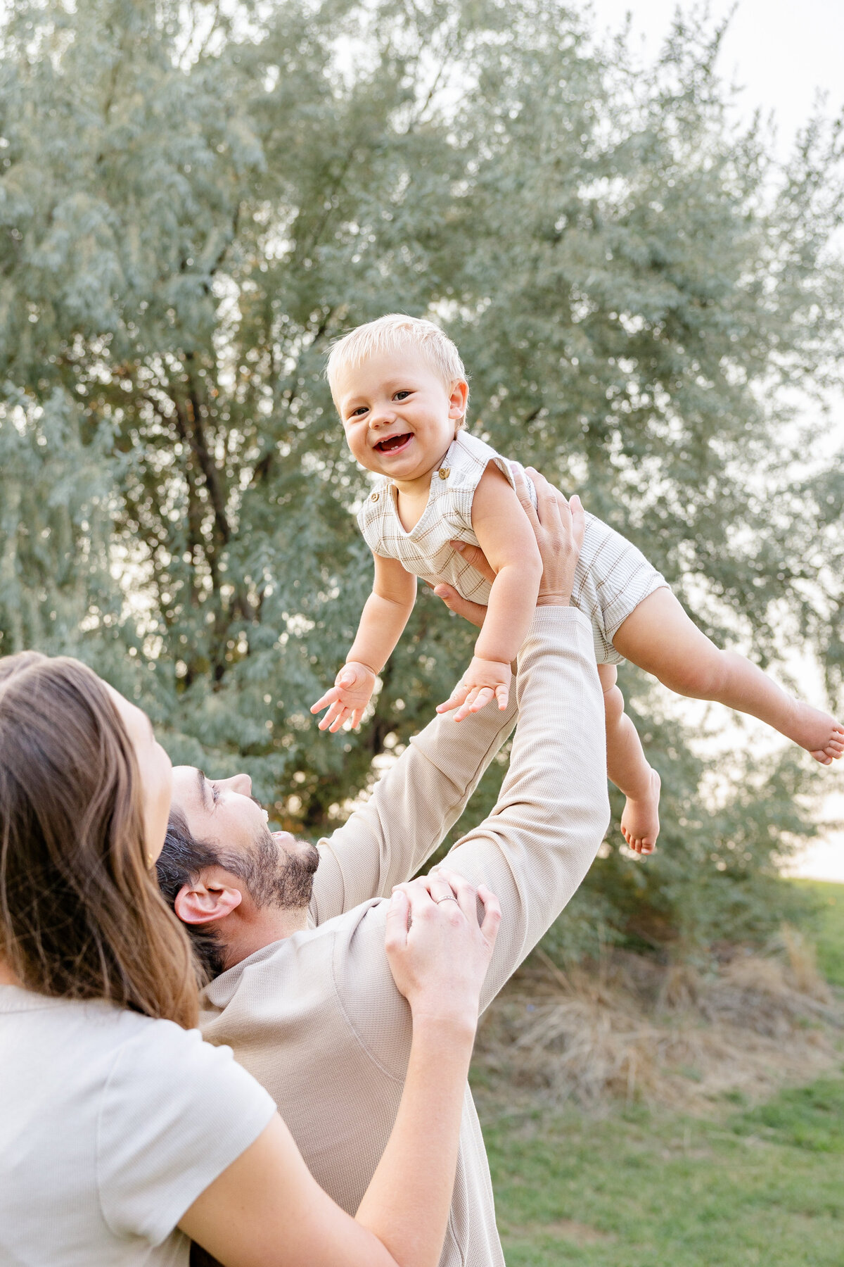 A mother and father lift their baby boy into the air, surrounded by lush greenery in Eagle Mountain, Utah. The baby's laughter and bright eyes radiate happiness.