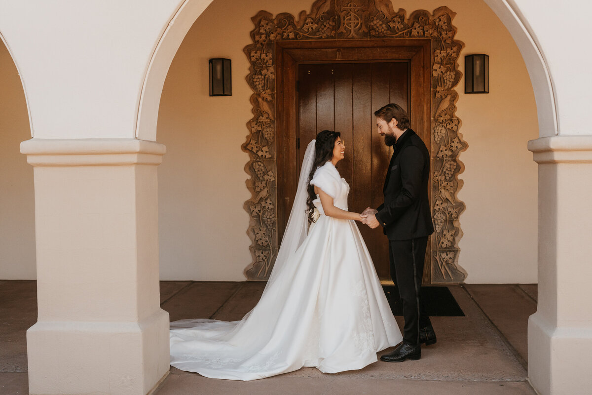 A wedding couple standing in an alcove holding hands and smiling 