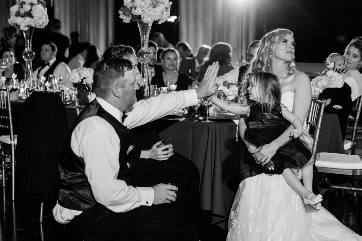 Candid reception moment at the Hall of State: groom Brad dancing with his mom while bride Emily holds the flower girl on her lap as she gives a high five to her dad, one of the groomsmen