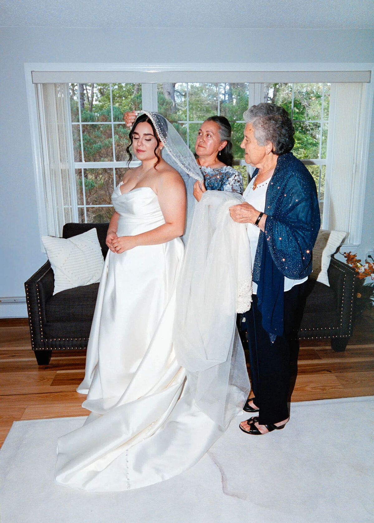 A bride in a white gown stands indoors while two older women help arrange her veil. Captured by a talented NJ wedding photographer, they all smile in a sunlit room with large windows, a dark couch, and hardwood floors.