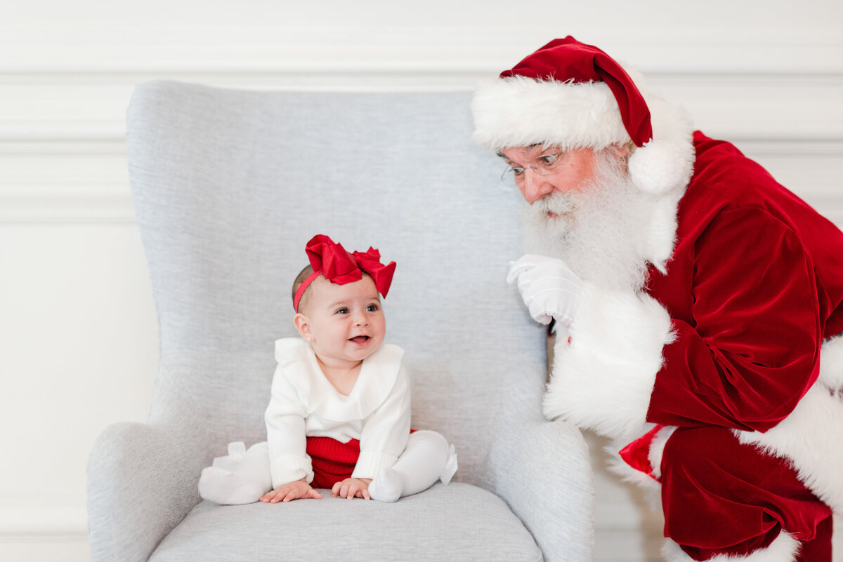 santa peeking over a baby girl sitting in a chair toddler siblings brother and sister jumping on the bed with snow and pajamas by miami christmas mini session photographer msp