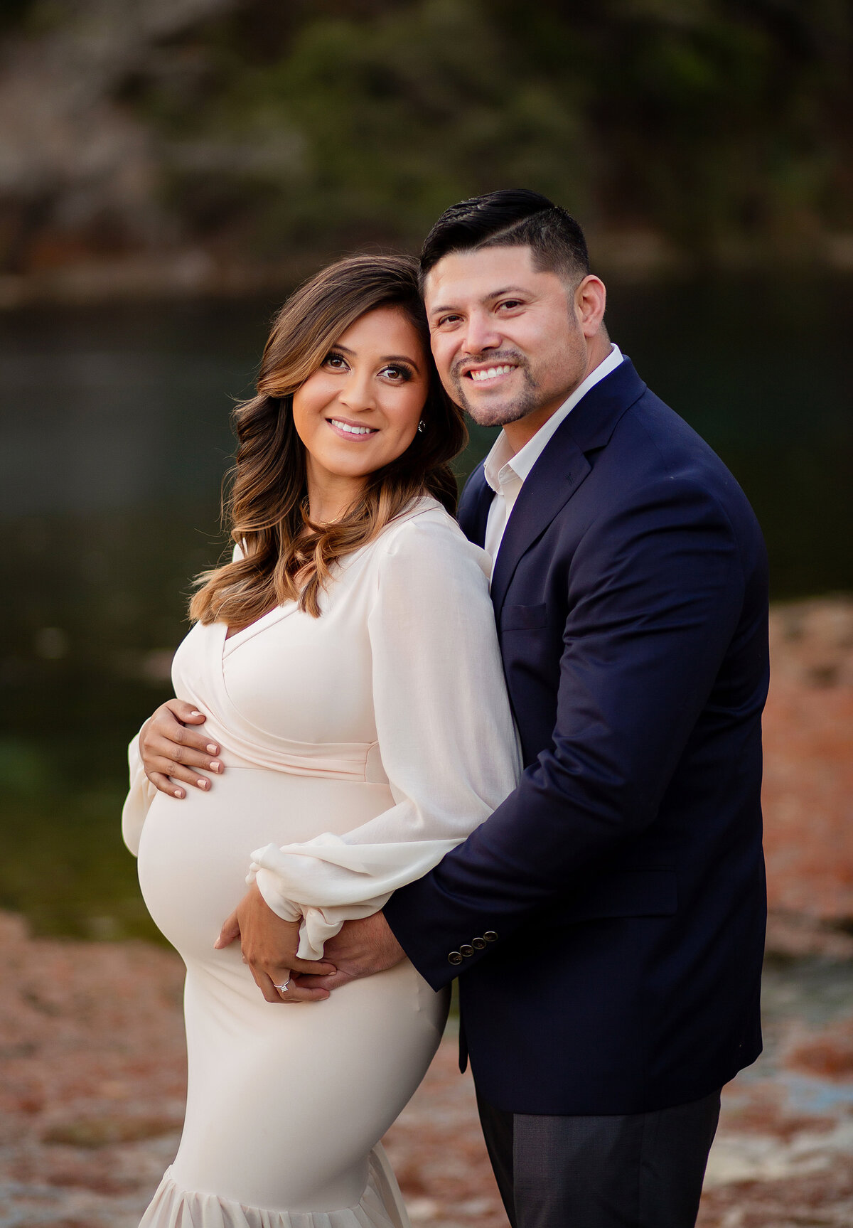 Mom and dad hold mom's pregnant belly while smiling at the camera in front of Fall Foliage