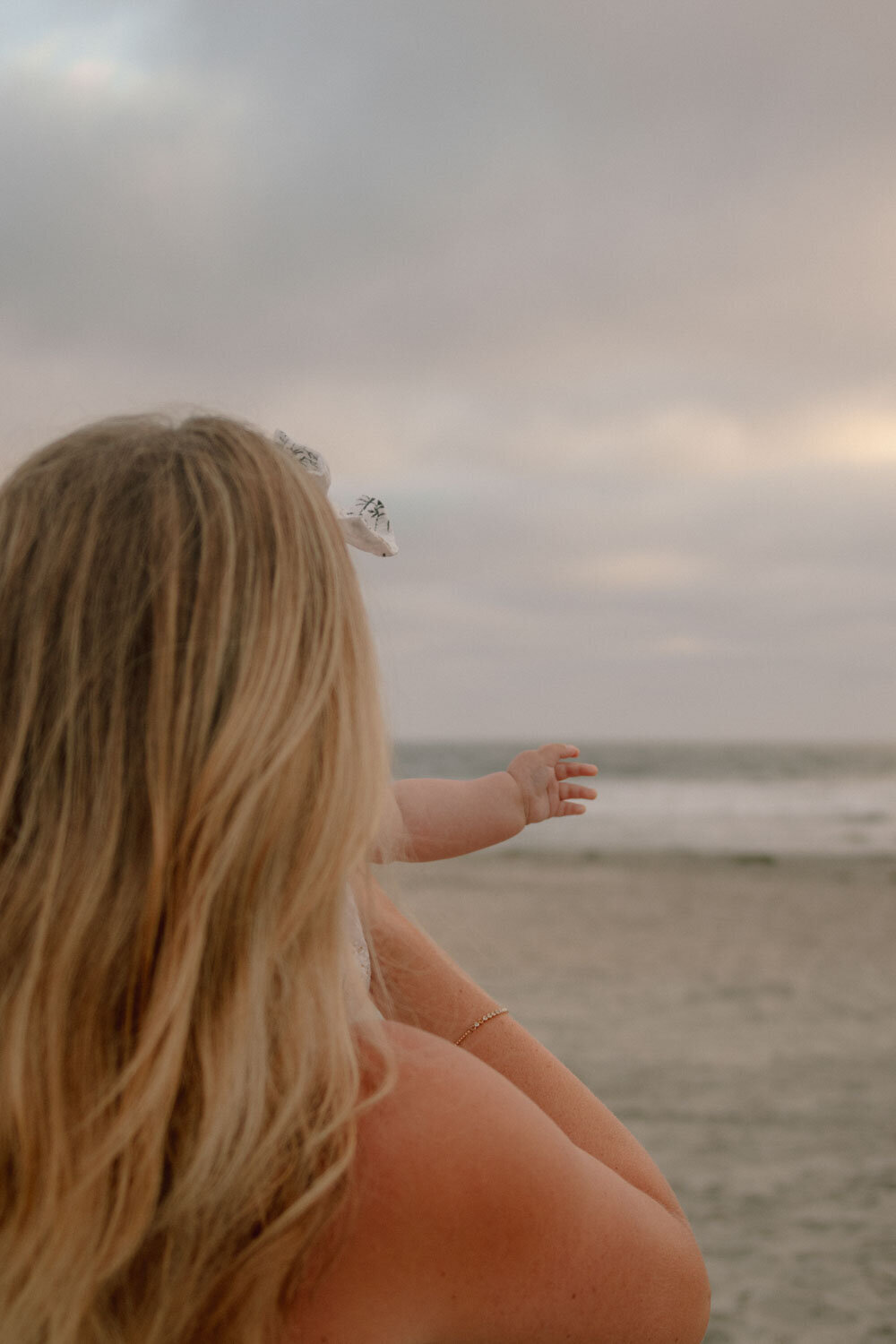 Mother holding her baby while standing on the beach, looking toward the ocean