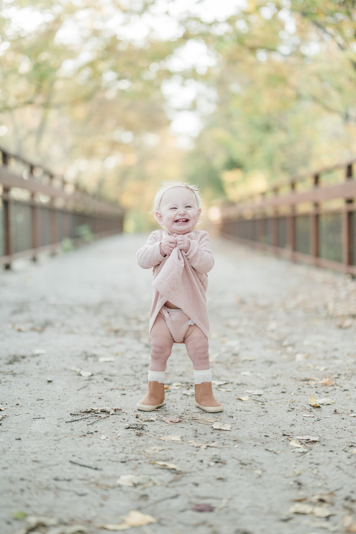 Happy baby girl standing on a leaf-covered wooden bridge wearing a blush pink outfit.
