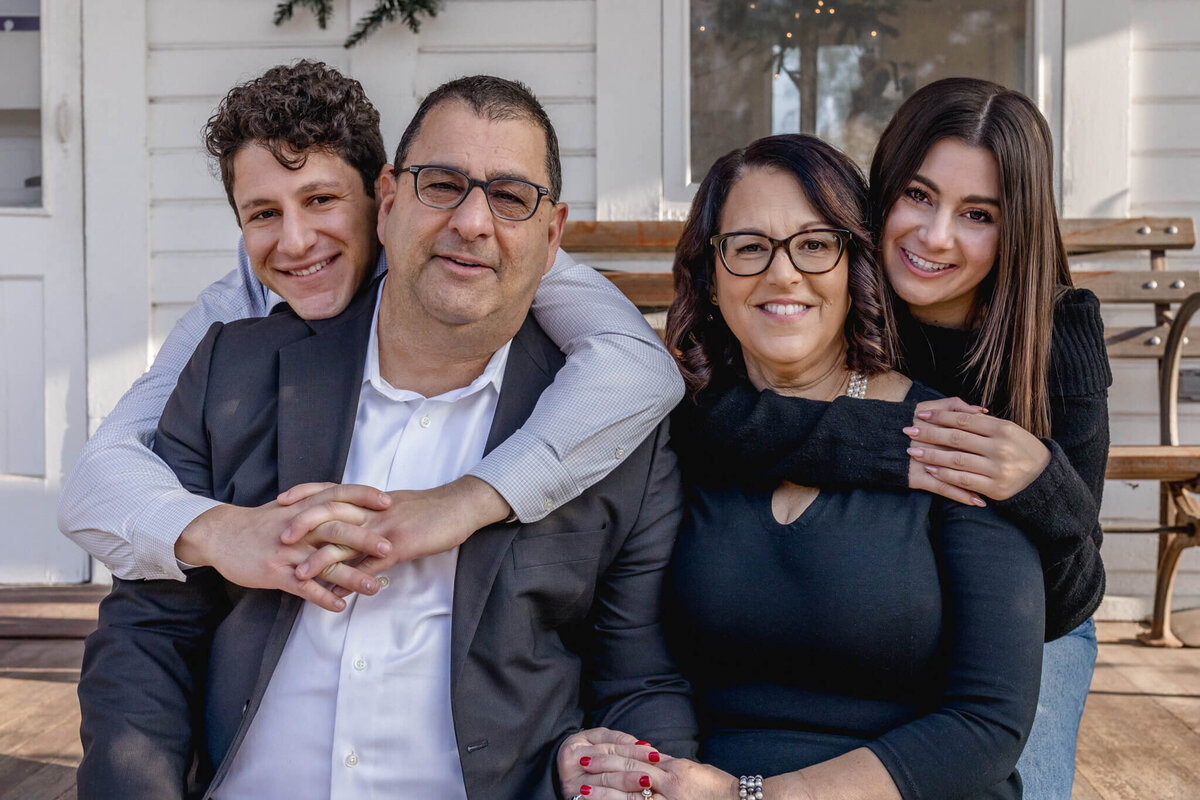 A husband and wife sitting next to each other and smiling at the camera while their son and daughter hug them from behind, resting their heads on their shoulders and smiling at the camera.