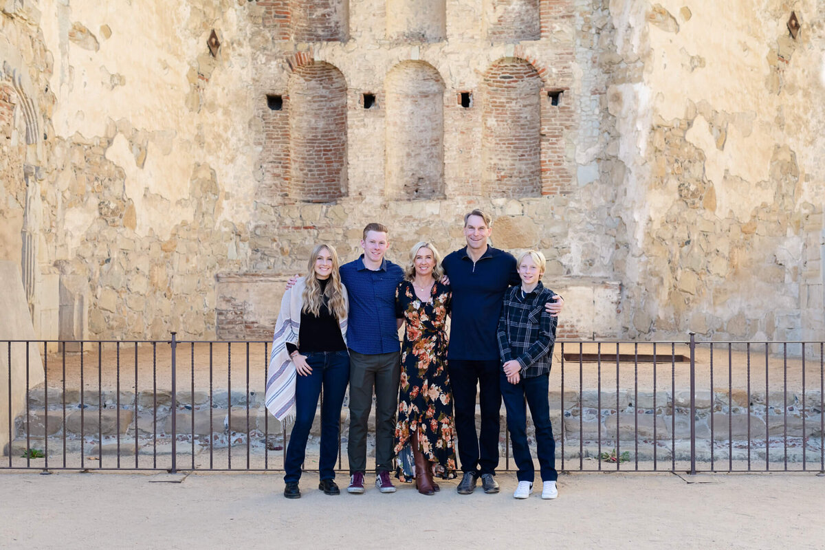 A blonde family of five standing in front of a black fence and a historic brick wall at Mission San Juan Capistrano.
