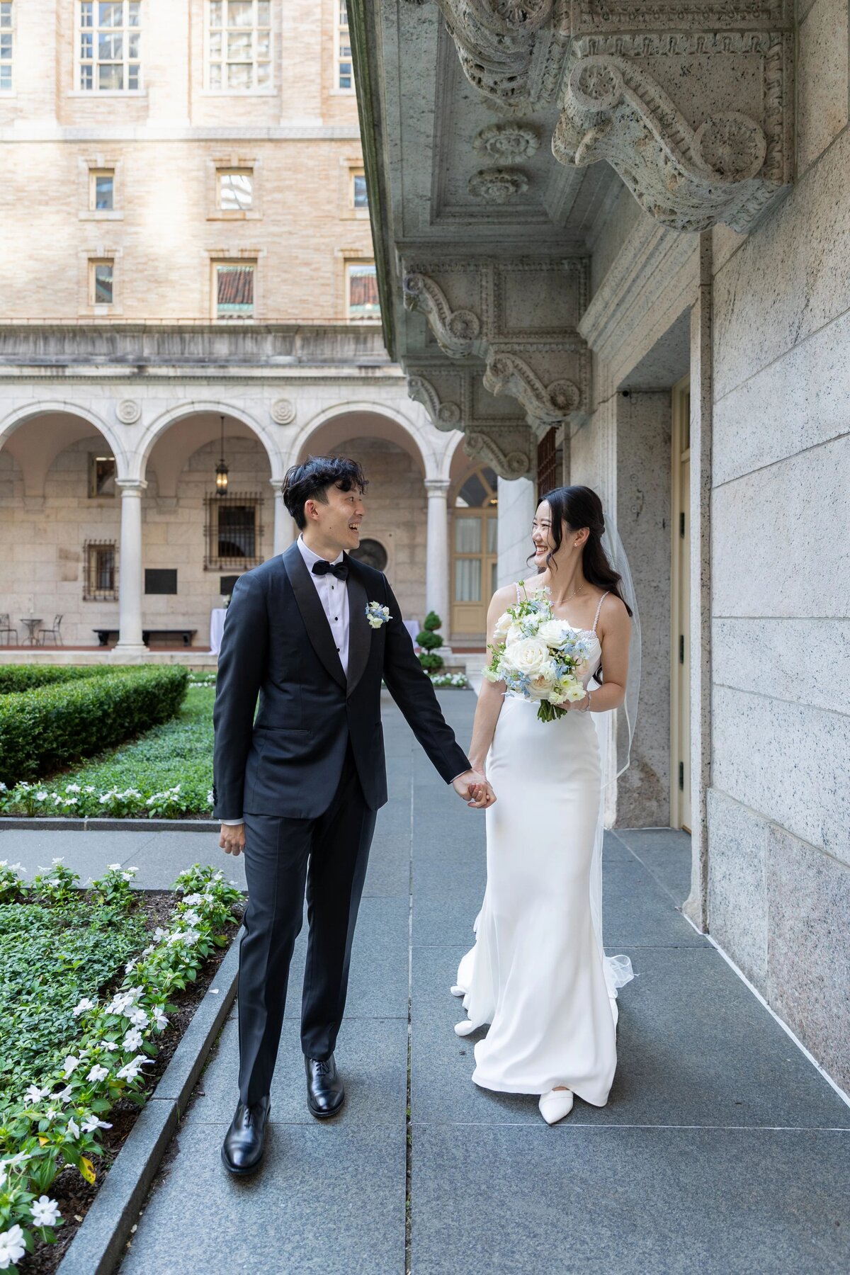 Wedding Photographers in RI | Kelsey Sheehan Photography Timeless Rhode Island Weddings | A bride in a white dress and a groom in a black tuxedo hold hands, smiling at each other in a courtyard with elegant arches and greenery.