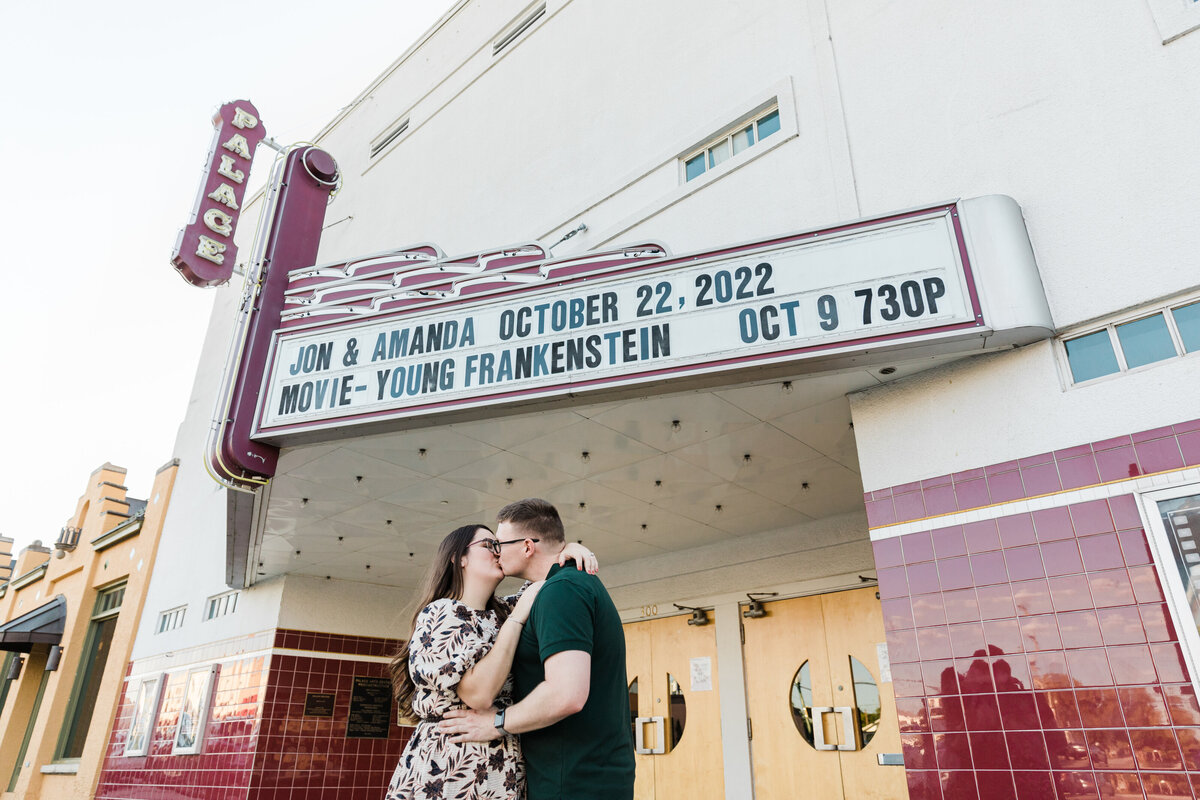 A couple sharing a kiss outside of a historic movie theater during their engagement session in downtown Grapevine, Texas. The woman on the left is wearing a white dress covered in black flowers and glasses. The man on the right is wearing a green shirt and glasses. The couple's name and wedding date is written above on the theater marquee.